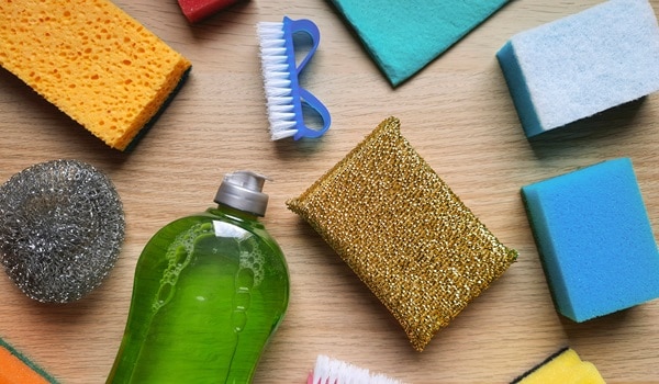 Kitchen sponges, scrubbing brushes, metal dishwashing sponge, rags, detergent on a wooden table. Variety of household cleaning products.
