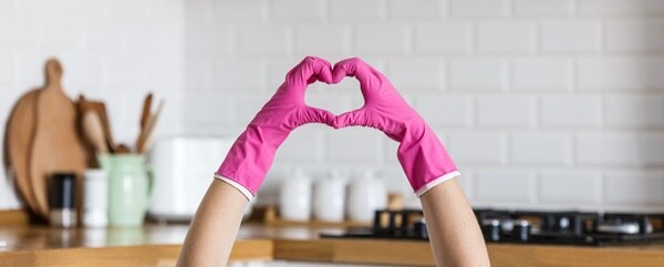 A woman's hands reach up from below the kitchen counter to form a heart with her pink protective gloves