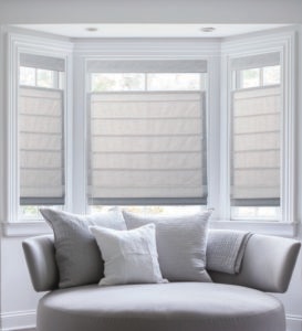 Large bay window with three white Roman shades, light filtering, in a bright living room with a white sofa and pillows