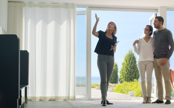 Professional Real Estate Agent Shows Stylish Modern House to a Beautiful Young Couple Who are in the Market for Purchasing/ Renting New Home. House Has Floor to Ceiling Windows and Seaside View.