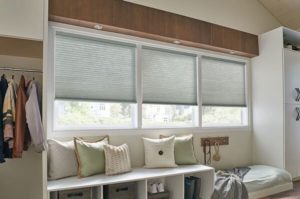 Light gray cellular shade covering a large window in a bedroom, with a white bench and pillows below